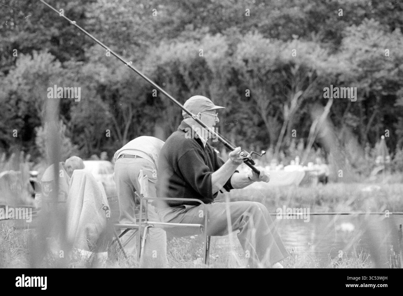 Gare di pesca IJmuiden, IJmuiden, Paesi Bassi, 19-08-1994 Whizgle News, Dutch Desk, i Paesi Bassi, 1950-2000 due persone praticano la pesca in prossimità di un sereno specchio d'acqua, uno seduto con una canna da pesca, mentre l'altro sembra prepararsi per il loro turno. La vegetazione lussureggiante li circonda, aggiungendovi una tranquilla atmosfera all'aperto. Foto Stock