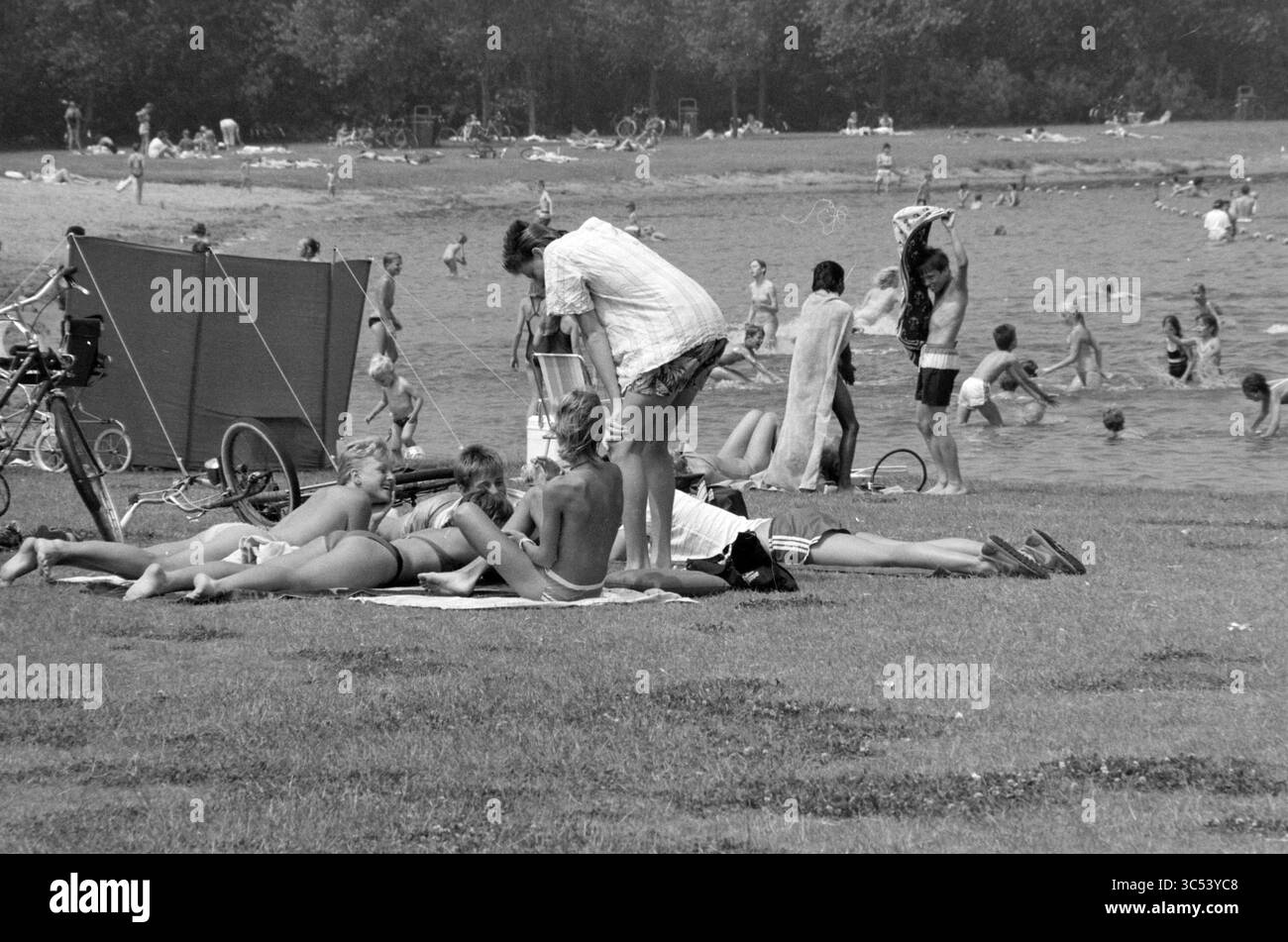 Folla estiva in un lago ricreativo, 26-06-1986 Whizgle News, Dutch Desk, Paesi Bassi, 1950-2000 Una vivace scena in una spiaggia dove gruppi di persone si rilassano sugli asciugamani e si godono il sole, mentre altri giocano in acqua o si dedicano a varie attività nelle vicinanze. Le biciclette sono parcheggiate, a indicare un'atmosfera rilassata e spensierata. Foto Stock