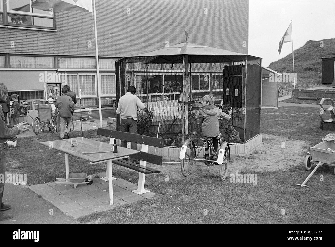 Playground Heliomare, giochi e giocattoli, 25-05-1971 Whizgle News, Dutch Desk, Paesi Bassi, 1950-2000 Un gruppo di persone partecipa a varie attività nei pressi di un recinto recintato, con bambini che giocano e adulti che interagiscono. Nelle vicinanze, una panchina e un tavolo sono posizionati sull'erba, suggerendo un rilassante ritrovo all'aperto. Le bandiere sventolano sullo sfondo, aggiungendo un'atmosfera vivace alla scena. Foto Stock