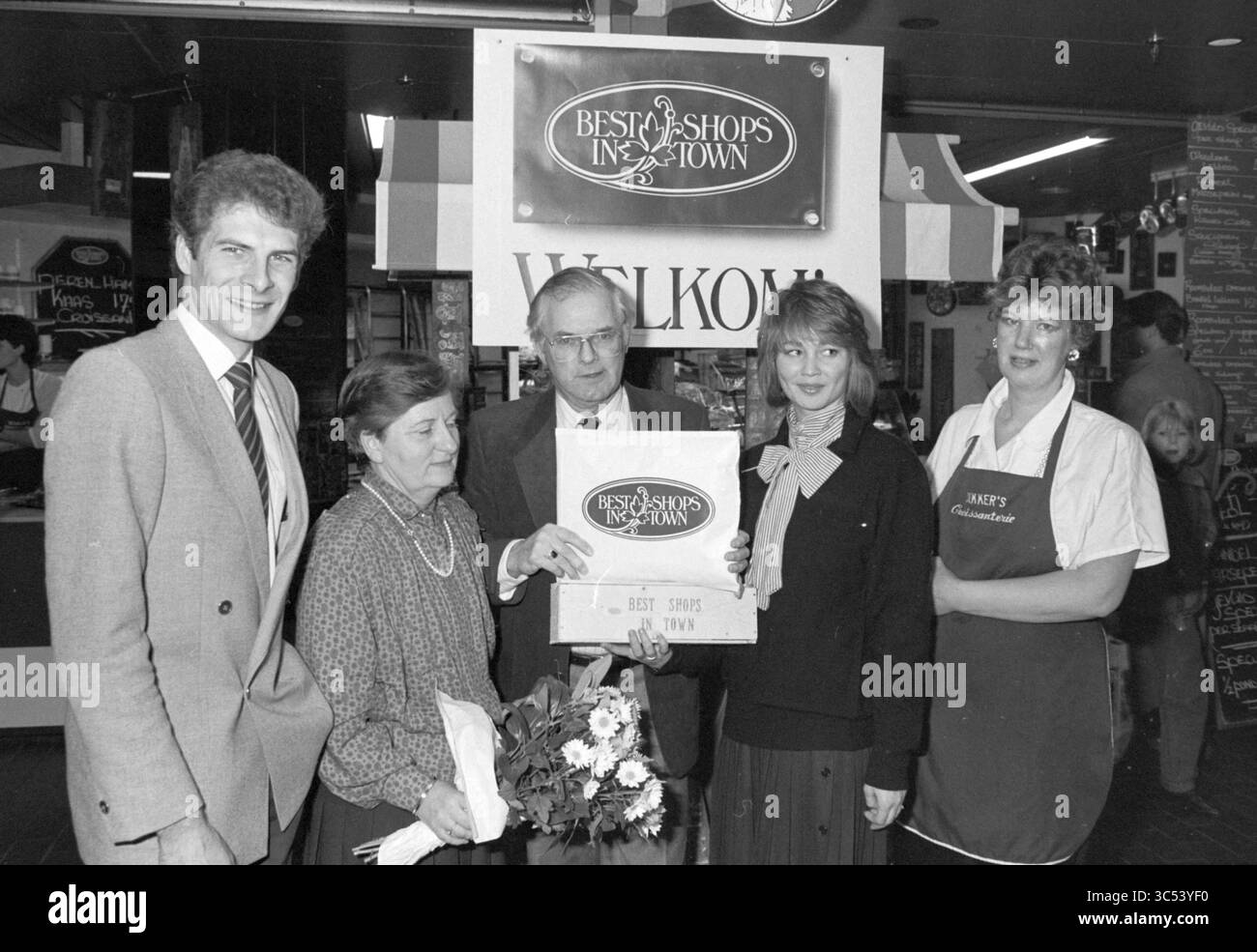 Cerimonia di premiazione per il miglior negozio in città, cerimonia di premiazione, 04-12-1985 Whizgle News, Dutch Desk, Paesi Bassi, 1950-2000 Un gruppo di cinque persone è orgoglioso di trovarsi in un vivace mercato, tenendo in mano un cartello con la scritta "Best Shops in Town". La scena raffigura un momento di festa, con un individuo che presenta fiori, circondato da sorrisi e da una calda atmosfera di riconoscimento della comunità. Foto Stock