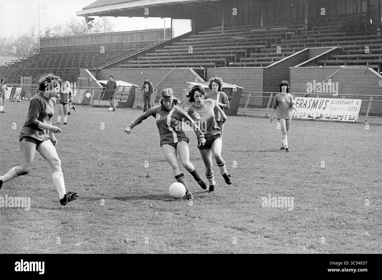 Calcio femminile. H'stede, Queen's Day, 01-05-1978 Whizgle News, Dutch Desk, Paesi Bassi, 1950-2000 Una scena dinamica cattura una partita di calcio femminile, con i giocatori concentrati sul pallone e che mostrano il loro atletismo. Sullo sfondo, una tribuna vuota e striscioni promozionali suggeriscono l'ambientazione dell'evento, mentre le divise dei giocatori e le espressioni decise evidenziano il loro spirito competitivo. Foto Stock