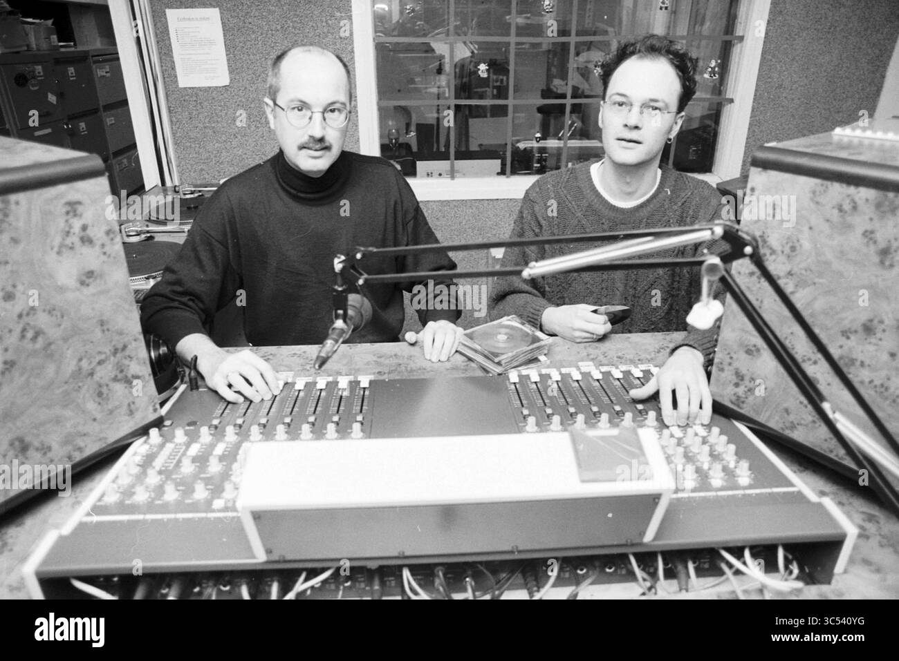 Renkema + Klepper, radio Logos, IJm., IJmuiden, Paesi Bassi, 18-12-1997 Whizgle News, Dutch Desk, Paesi Bassi, 1950-2000 due persone sono sedute ad una console audio in uno studio, impegnati in una conversazione. La sala è piena di apparecchiature audio e l'atmosfera suggerisce un ambiente creativo o di trasmissione. Una persona che indossa occhiali e un maglione guarda attentamente la console, mentre l'altra sembra controllare un dispositivo in mano. Foto Stock