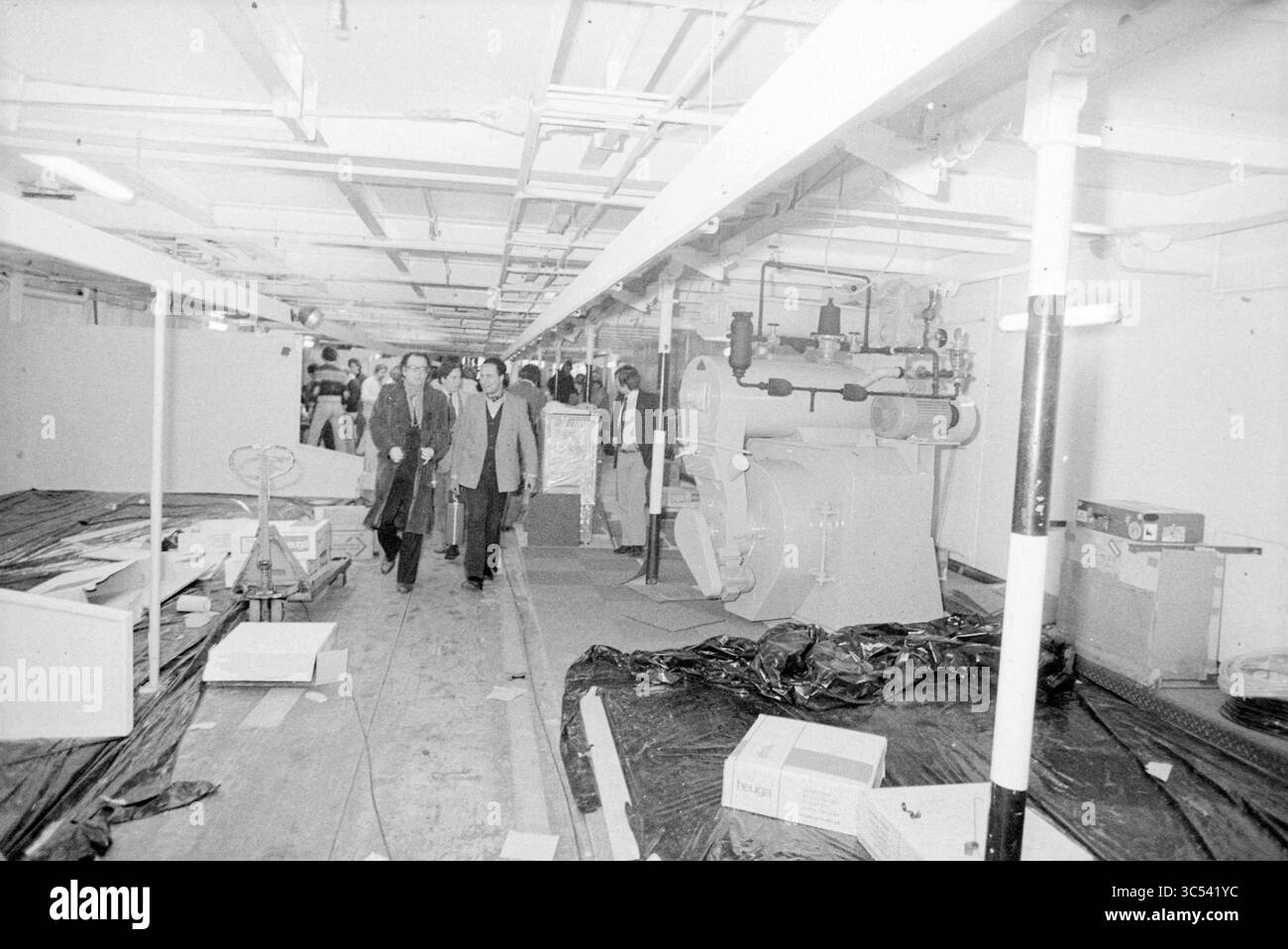 Holland Expo, Ships, 13-01-1979 Whizgle News, Dutch Desk, Paesi Bassi, 1950-2000 Un gruppo di persone attraversa un lungo corridoio industriale adornato di macchinari e materiali da costruzione, suggerendo un processo di ristrutturazione o ispezione in corso in un grande stabilimento. Foto Stock