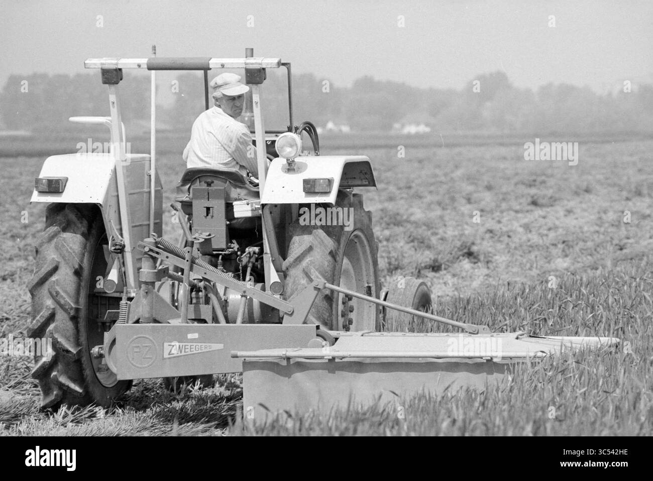 Aratura campo di grano, 31-05-1994 Whizgle News, Dutch Desk, Paesi Bassi, 1950-2000 Un agricoltore gestisce un trattore in un vasto campo, utilizzando abilmente attrezzature per la cura delle colture, circondato da una vegetazione lussureggiante e da alberi distanti. Foto Stock