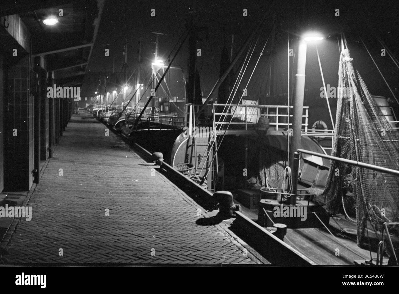 Navi nel porto di pesca di sera, navi che scaricano pesce, cernita, pesca e altri, pescivendoli, pesca, 04-12-1963 Whizgle News, Dutch Desk, Paesi Bassi, 1950-2000 Una tranquilla scena sul lungomare illuminata da lampioni stradali, dove le barche da pesca sono ormeggiate lungo un sentiero acciottolato, creando un'atmosfera serena e riflettente. Foto Stock