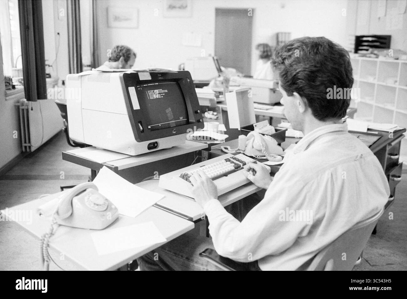 Uomo dietro lo schermo del computer, reparto di distribuzione delle abitazioni, computer, carenza di alloggi e alloggi, 03-06-1986 Whizgle News, Dutch Desk, Paesi Bassi, 1950-2000 Una persona siede a una scrivania scrivendo su un computer, circondato da documenti e un telefono vecchio stile, mentre altri individui lavorano alle scrivanie adiacenti in un ambiente di ufficio spazioso. Foto Stock
