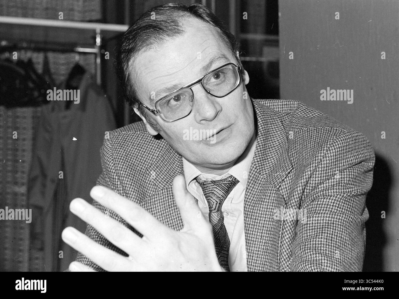 Dinkelberg RCH., Football R.C.H., 17-12-1980 Whizgle News, Dutch Desk, Paesi Bassi, 1950-2000 Un uomo in costume e occhiali si siede a un tavolo, gestendo con la mano mentre si mette in conversazione. La sua espressione è premurosa e riflette una discussione seria. Sullo sfondo, l'abbigliamento su grucce suggerisce un ambiente professionale. Foto Stock