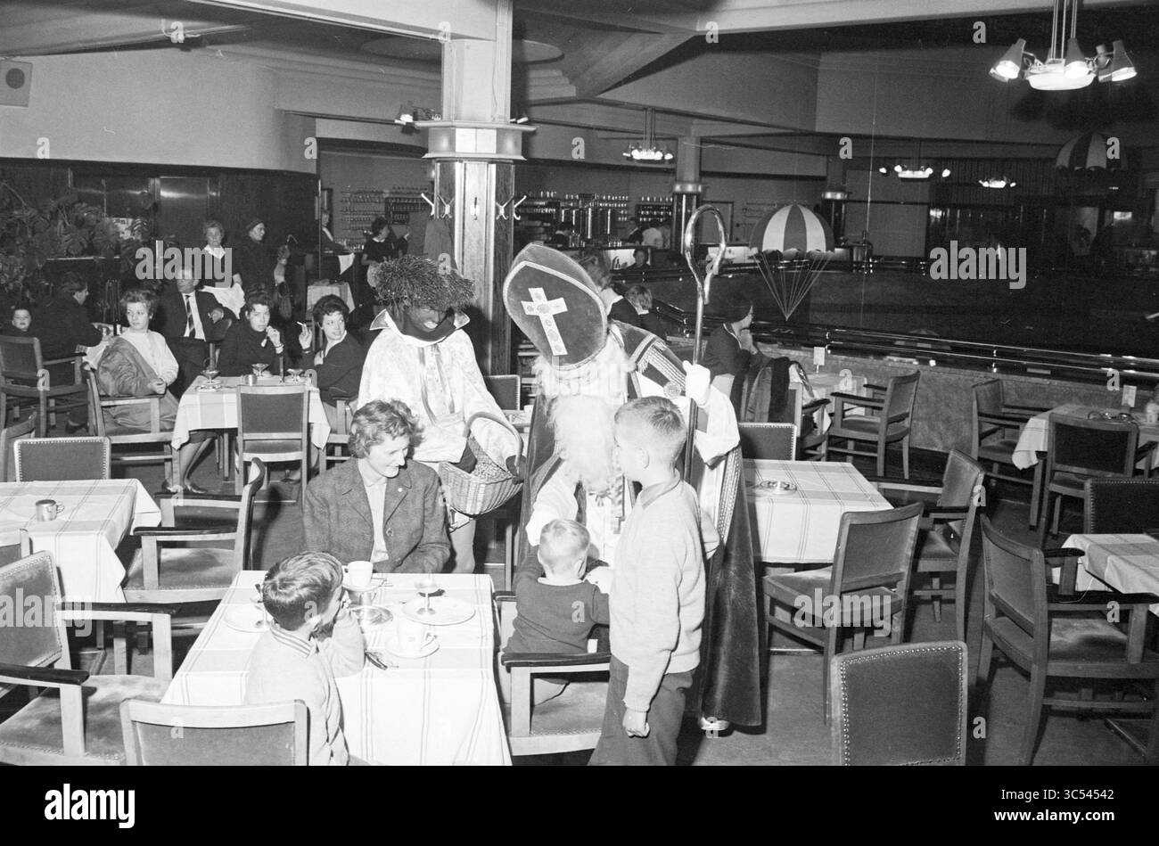 Saint Nicholas at Vroom and Dreesmann Lunchroom, St. Nicholas, Sinterklaas, 18-11-1963 Whizgle News, Dutch Desk, i Paesi Bassi, 1950-2000, Una vivace scena di ristoranti in cui un gruppo di persone, tra cui persone in costumi tradizionali, interagisce con una coppia di bambini seduti a un tavolo. I ristoranti nelle vicinanze osservano l'atmosfera festosa, che include decorazioni natalizie e un'aria di festa. Foto Stock