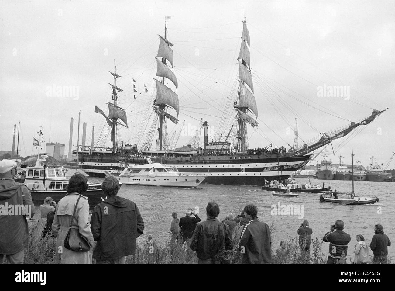 Arrival Sail 1980, IJmuiden, Sailing, IJmuiden, Paesi Bassi, 06-08-1980 Whizgle News, Dutch Desk, Paesi Bassi, 1950-2000 Una grande nave alta con vele fluttuanti scivola attraverso l'acqua, accompagnata da barche più piccole mentre gli osservatori si radunano lungo la riva, affascinati dallo spettacolo marittimo. Sullo sfondo, gli edifici industriali e i montanti suggeriscono l'ambientazione del porto. Foto Stock