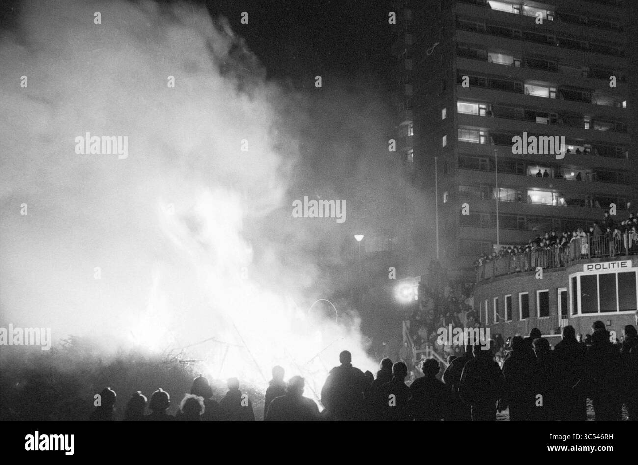 Fuoco sull'albero di Natale sulla spiaggia di Zandvoort, alberi di Natale e di Natale, Zandvoort, 03-01-1990 Whizgle News, Dutch Desk, Paesi Bassi, 1950-2000 Una folla si raduna in primo piano, sagomata contro le fiamme e il fumo che sale da un incendio, mentre un edificio si infila sullo sfondo, con gli spettatori che osservano dai piani più alti. I servizi di emergenza sono visibili nelle vicinanze, a indicare una situazione di tensione che si sta sviluppando. Foto Stock