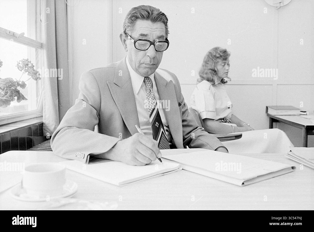 Ritratto di un uomo Whizgle News, Dutch Desk, Paesi Bassi, 1950-2000 Un uomo in abito formale, con occhiali, concentrato a prendere appunti in un quaderno, mentre una donna si siede sullo sfondo, osservando tranquillamente a un tavolo adornato da una tazza. La luce soffusa viene filtrata attraverso una finestra, evidenziando l'atmosfera semplice e professionale. Foto Stock