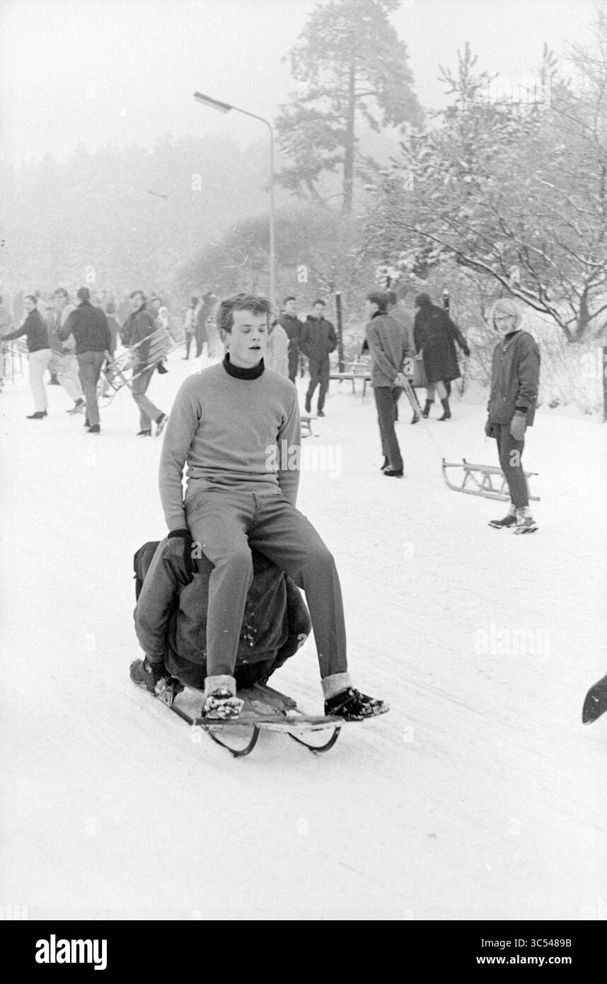 Attività e divertimento sulla neve negli anni '1960 Whizgle News, Dutch Desk, Paesi Bassi, 1950-2000 Un giovane uomo siede con sicurezza su una slitta mentre un amico lo tira lungo un sentiero innevato, mentre altri si divertono nelle vicinanze. La scena cattura lo spirito gioioso di una giornata innevata, con alberi spolverati di bianco e persone impegnate in varie attività. Foto Stock