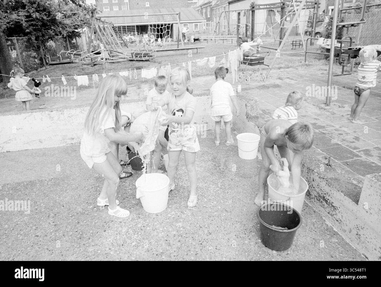 Il giorno del lavaggio delle bambole, il parco giochi dei soffiatori di vetro, bambini, bambini, case per bambini, festa per bambini, bambini, Haarlem, Scheepmakersdijk, Paesi Bassi, 13-07-1987 Whizgle News, Dutch Desk, Paesi Bassi, 1950-2000 Un gruppo di bambini gioca gioiosamente in un ambiente all'aperto, impegnandosi in un'attività acquatica. Alcuni bambini riempiono secchi e spruzzano acqua, mentre altri sono occupati con vari giocattoli e attività nelle vicinanze. I vestiti sono appesi a una fila e le attrezzature per il parco giochi sono visibili sullo sfondo, creando un'atmosfera vivace e spensierata. Foto Stock