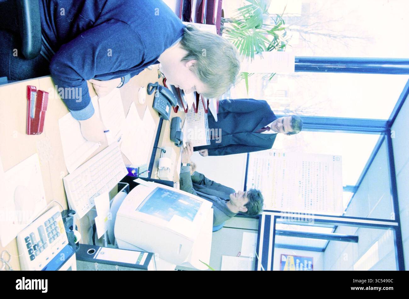Varie, reparto + personale + gruppo, 31-01-2001 Whizgle News, Dutch Desk, Paesi Bassi, 1950-2000 Un ambiente d'ufficio molto frequentato con tre persone impegnate in discussioni e attività lavorative. Una persona è concentrata su un computer, mentre un'altra si appoggia sulla scrivania per rivedere i documenti. Un terzo individuo si trova nelle vicinanze, contribuendo alla conversazione. Lo spazio di lavoro è organizzato con varie forniture per ufficio e una grande finestra che consente di illuminare l'ambiente circostante con luce naturale. Foto Stock