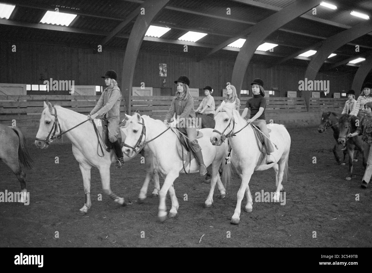 Giovani che cavalcano a cavallo in un'arena coperta di una scuola di equitazione. Whizgle News, Dutch Desk, The Netherlands, 1950-2000 un gruppo di cavalieri a cavallo naviga in una spaziosa arena al coperto, mostrando un mix di espressioni mirate e cameratismo. Ogni pilota è comodamente seduto sui propri cavalli, con una serie di equipaggiamento da equitazione e caschi. Intorno a loro ci sono altri cavalli e persone, che si aggiungono alla vivace atmosfera dell'attività equestre. Foto Stock