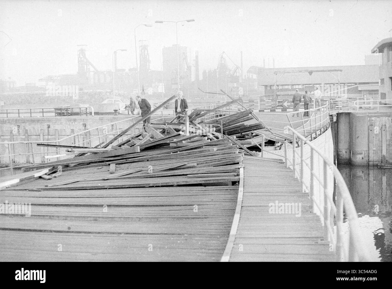 Chiusa in IJmuiden colpita dalla nave, IJmuiden, Nederland Whizgle News, Dutch Desk, Paesi Bassi, 1950-2000 due persone navigano su un ponte di legno danneggiato, con detriti sparsi attraverso il passaggio pedonale. Sullo sfondo si insinuano strutture industriali che fanno pensare ad un'area un tempo animata di fronte al mare. Foto Stock