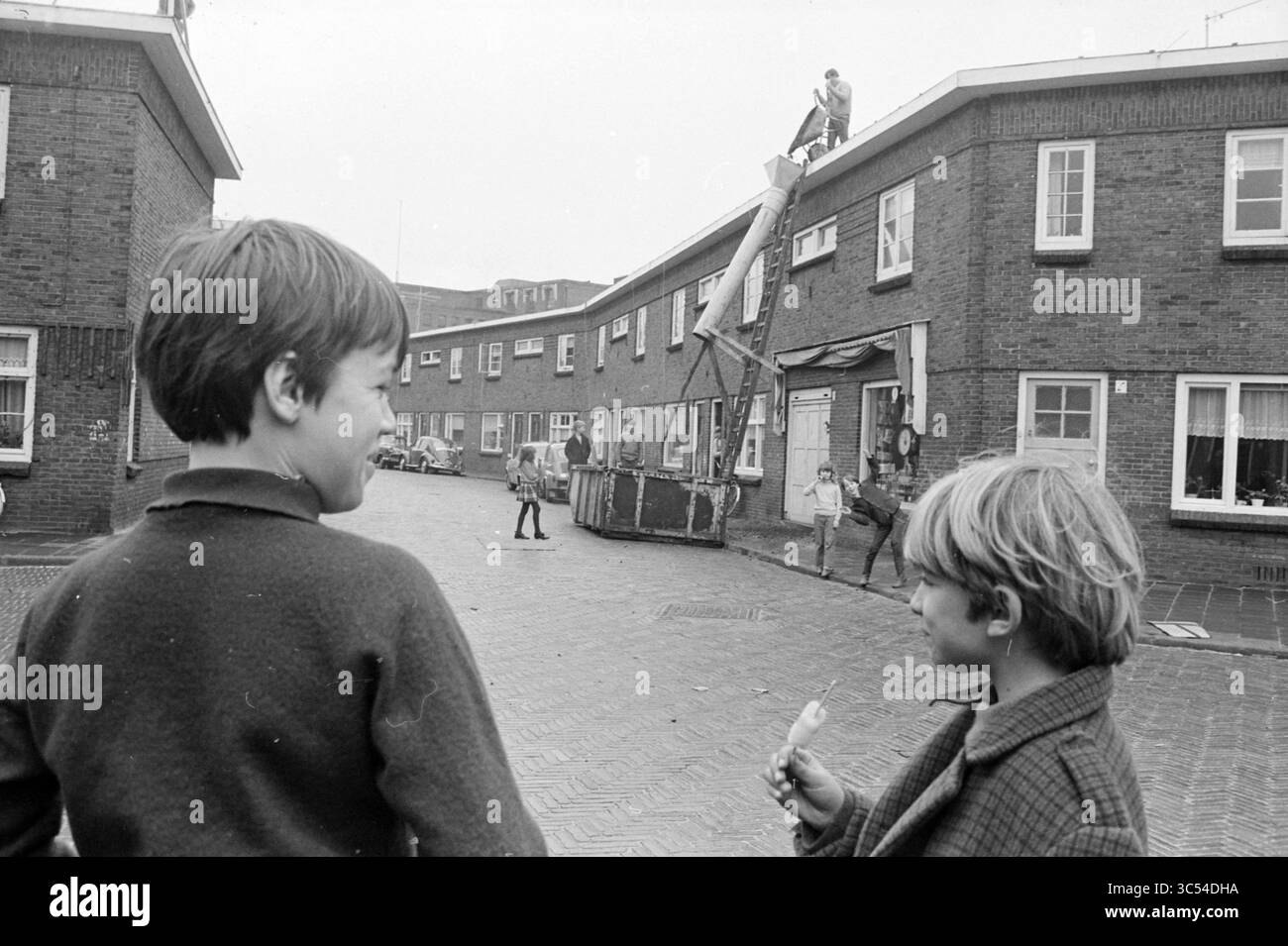 Thorbecke Neighborhood, Streets, 04-02-1970 Whizgle News, Dutch Desk, Paesi Bassi, 1950-2000 due ragazzi stanno in un cortile residenziale, sorridendo e chiacchierando mentre guardano un gruppo di bambini impegnati in attività ludiche nelle vicinanze. Un ragazzo tiene uno spuntino, mentre un altro osserva una persona sul tetto, suggerendo un vivace pomeriggio di esplorazione e divertimento nel loro quartiere. Foto Stock