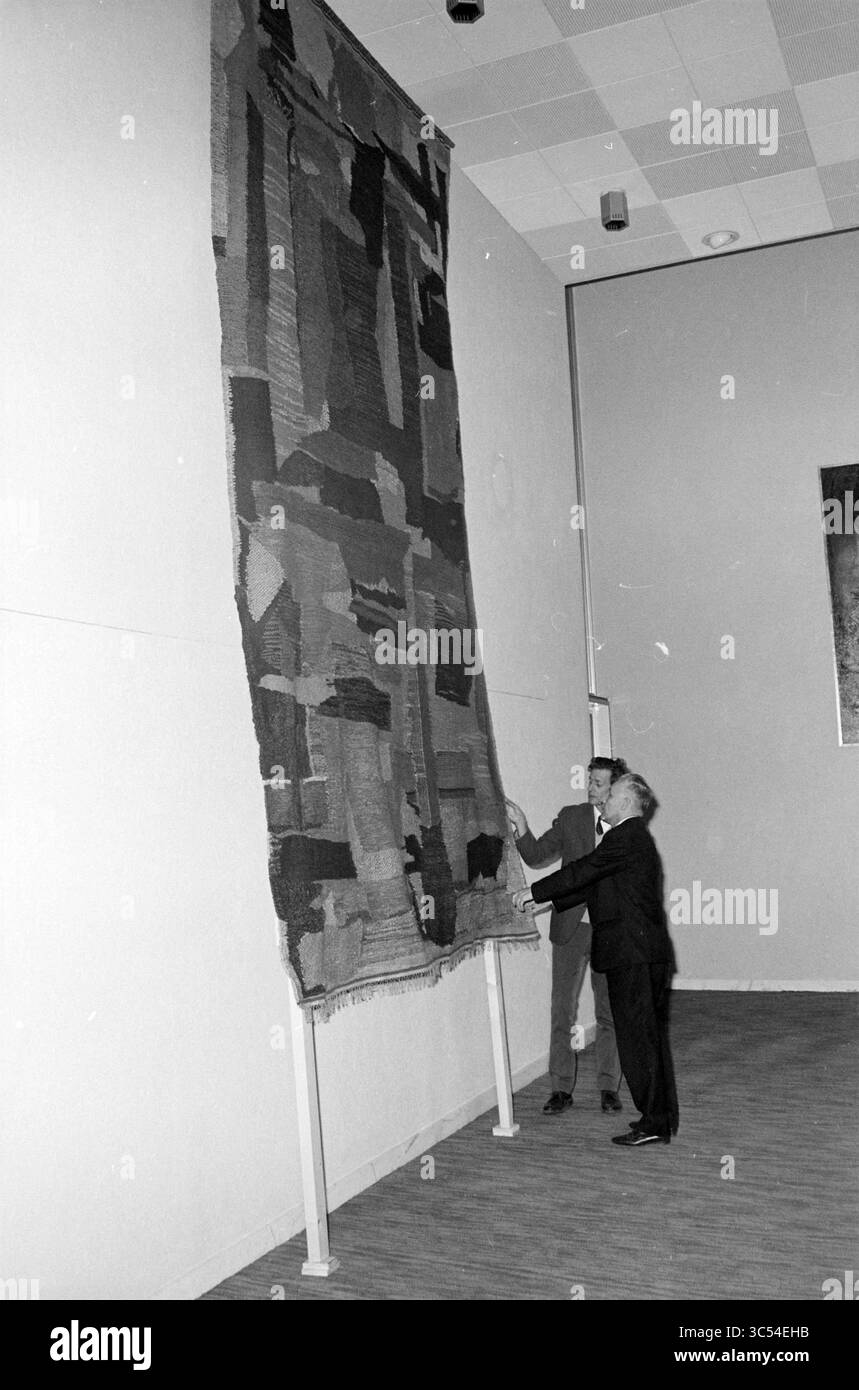 Alcuni uomini guardano un grande arazzo Whizgle News, Dutch Desk, Paesi Bassi, 1950-2000 due individui ispezionano un'opera tessile astratta montata su un muro. L'arazzo presenta una varietà di texture e motivi geometrici, che mostrano un mix di colori e forme. L'ambientazione sembra essere uno spazio espositivo, che enfatizza le dimensioni e la complessità artistica dell'opera. Foto Stock