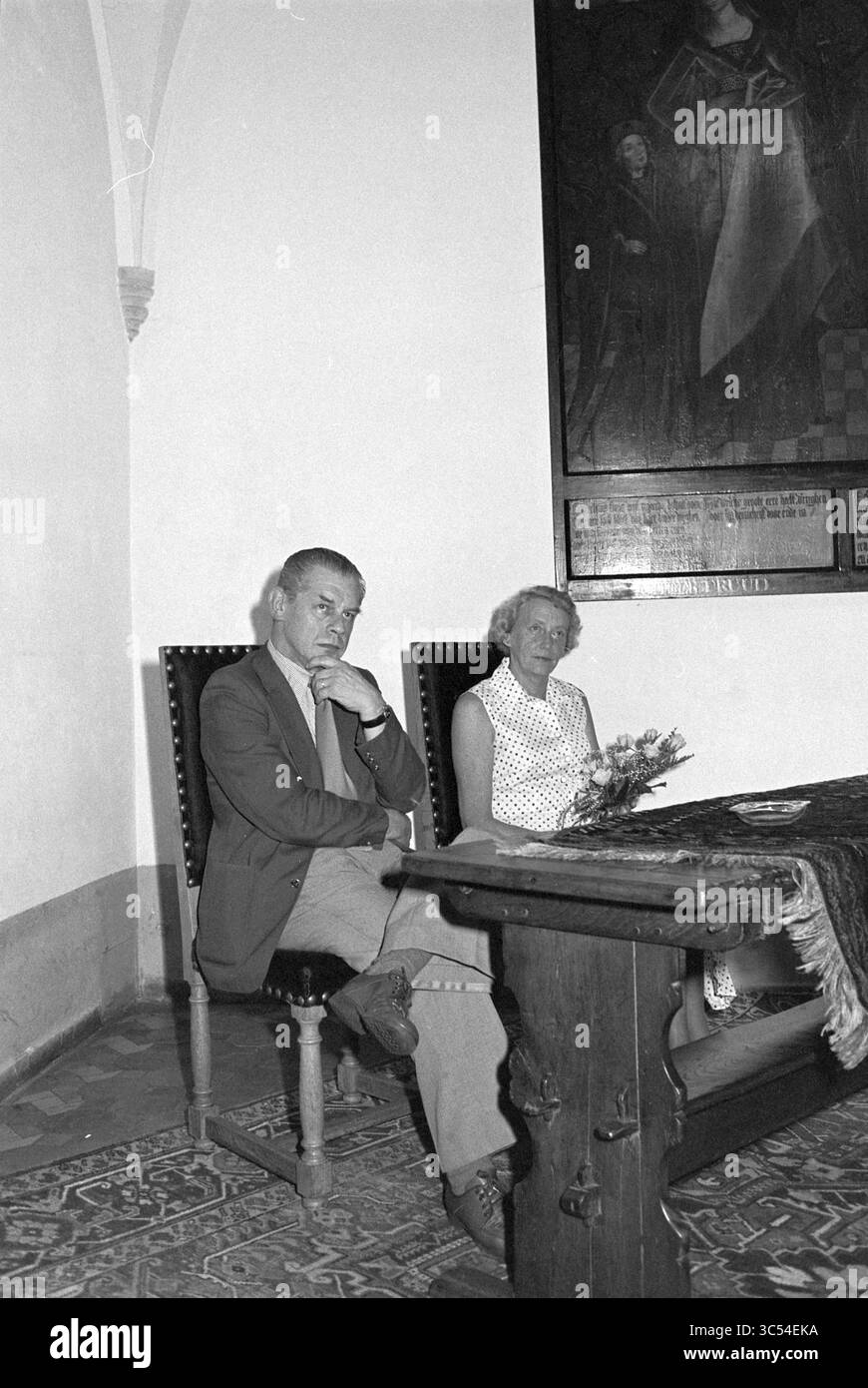 Lode Mr. Moens, Haarlem City Hall, Homage, 06-09-1977 Whizgle News, Dutch Desk, i Paesi Bassi, 1950-2000 Un uomo e una donna si siedono fianco a fianco a fianco a un tavolo di legno, nel profondo del pensiero. La donna regge un piccolo bouquet di fiori e sono circondate da un interno storico caratterizzato da decorazioni ornamentali e da un grande dipinto murale sullo sfondo. Foto Stock