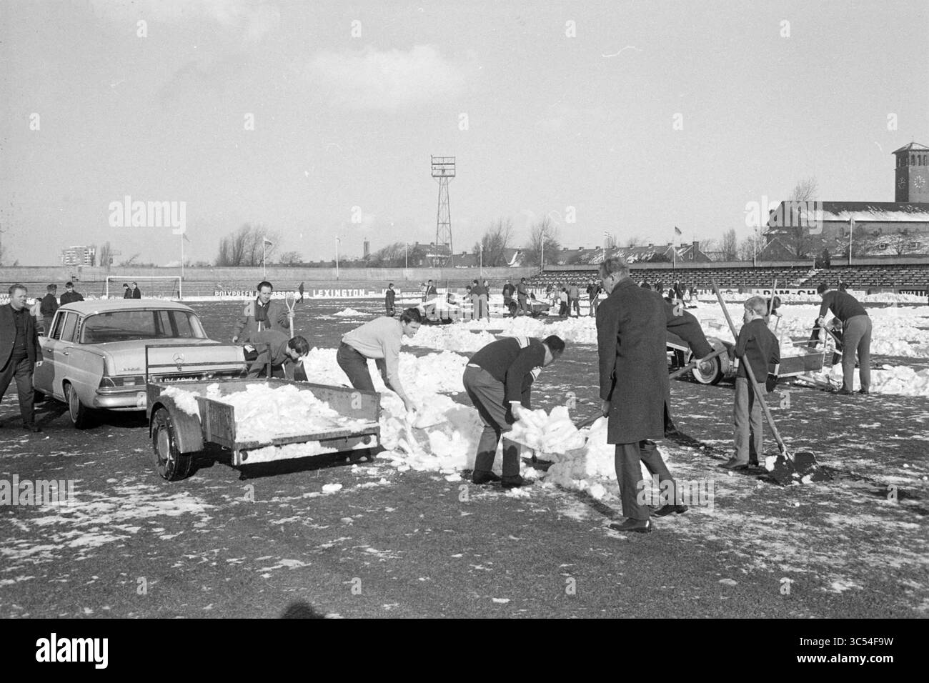 Clearing Snow in Haarlem Grounds, Football Haarlem, Work, 04-02-1968 Whizgle News, Dutch Desk, i Paesi Bassi, 1950-2000 Un gruppo di persone vestite di abbigliamento invernale sta sgombrando una zona innevata, utilizzando pale e rimorchi per trasportare la neve. Sullo sfondo, sono visibili strutture e una torre, mentre i veicoli parcheggiati sono visti nelle vicinanze, indicando uno sforzo organizzato per rimuovere la neve dal suolo. Foto Stock