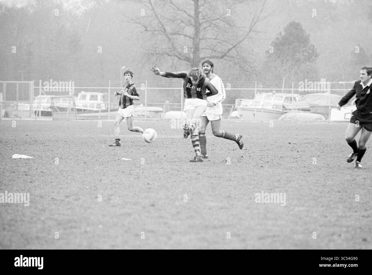 RCH - Zeeburgia, Football R.C.H., 09-03-1980 Whizgle News, Dutch Desk, Paesi Bassi, 1950-2000 Una vivace partita di calcio si svolge su un campo erboso, con i giocatori in azione. Un giocatore dribbla abilmente la palla mentre compagni di squadra e avversari navigano nel gioco. Sullo sfondo sono presenti alberi e barche parcheggiate, che aggiungono un'atmosfera unica alla scena. Foto Stock