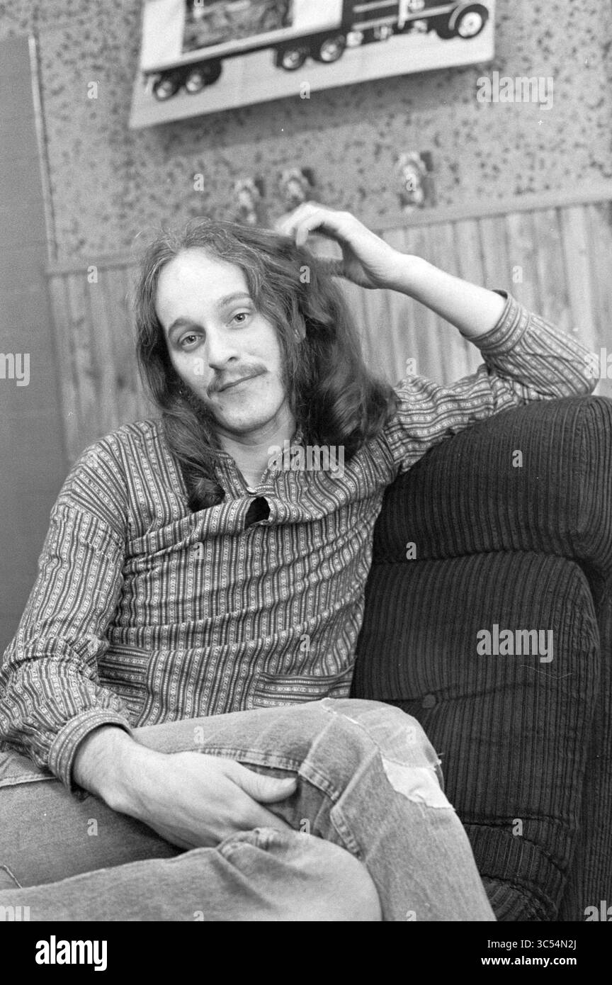 Ritratto Sido Martens, cantante del gruppo Fungus, Zang e cori, 10-11-1975 Whizgle News, Dutch Desk, Paesi Bassi, 1950-2000 Un giovane rilassato si siede su un divano, correndo casualmente le dita tra i suoi lunghi capelli, indossando una camicia a righe e jeans angosciati. Lo sfondo presenta una parete a motivi geometrici con elementi decorativi, che suggeriscono uno spazio accogliente e vissuto. Foto Stock