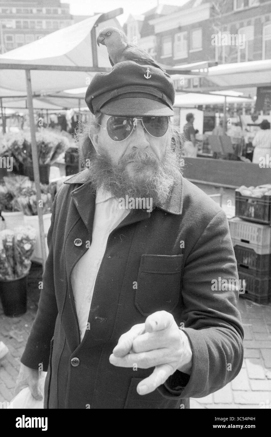 Uomo con pappagallo in testa, uccelli, 03-05-1986 Whizgle News, Dutch Desk, Paesi Bassi, 1950-2000 Un uomo barbuto che indossa occhiali da sole e un cappello da capitano si erge con sicurezza, puntando con una mano. Un piccolo pappagallo appollaiato sul cappello, aggiungendo un tocco giocoso al suo aspetto aspro, mentre le vivaci bancarelle del mercato e gli acquirenti incorniciano lo sfondo. Foto Stock