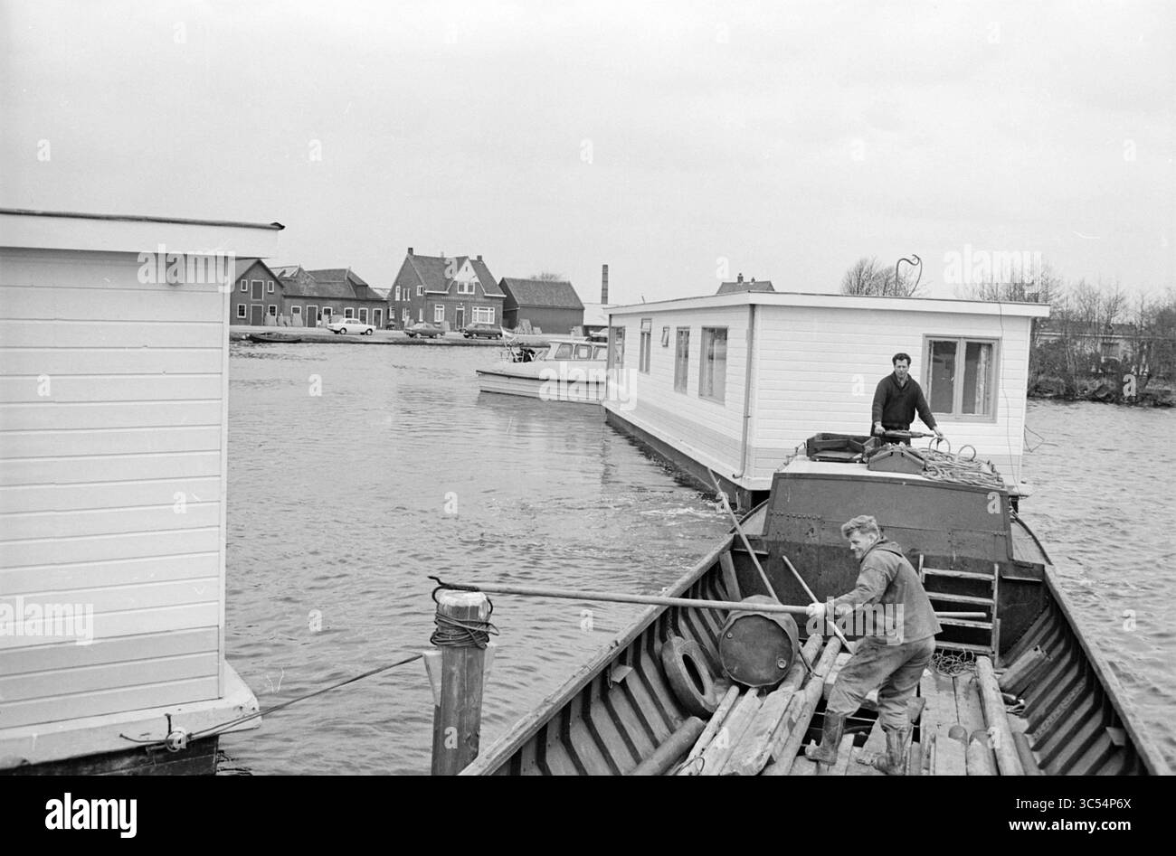 Trainando la casa galleggiante attraverso il Ringvaart in una nuova posizione, Aalsmeerderdijk Whizgle News, Dutch Desk, Paesi Bassi, 1950-2000 Un uomo lavora su una chiatta, usando un palo per navigare vicino a case galleggianti lungo un tranquillo corso d'acqua, mentre un altro individuo tende a una nave vicina sullo sfondo. La scena cattura la tranquillità della vita sull'acqua e l'architettura unica delle case galleggianti. Foto Stock