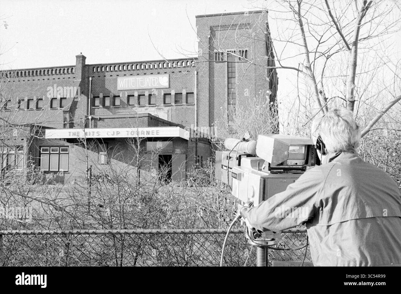 Registrazioni TV CJP in Witte Tejater IJmuiden, televisione, IJmuiden, Paesi Bassi, 14-11-1983 Whizgle News, Dutch Desk, Paesi Bassi, 1950-2000 Un cameraman si prepara a registrare all'esterno di un edificio in mattoni con cartelli che riportano "MITCHELL" e "THE MATS CJP TOURNEE", circondati da vegetazione e alberi sovradimensionati, indicando un evento o un luogo potenzialmente significativo. Foto Stock