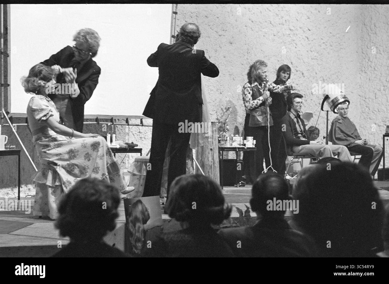 Dimostrazione di parrucchiere a Schalkererf, dimostrazione, parrucchieri e parrucchieri, Haarlem, Bernadottelaan, Paesi Bassi, 24-02-1975 Whizgle News, Dutch Desk, Paesi Bassi, 1950-2000 sul palco si sta svolgendo una dimostrazione di parrucchiere dal vivo, con stilisti che mostrano le loro tecniche sui modelli. Il pubblico guarda attentamente, mentre vari strumenti e prodotti per lo styling sono disposti nelle vicinanze. L'atmosfera è vivace e riflette un mix di creatività e performance. Foto Stock