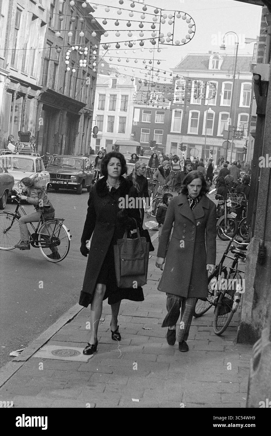 Gente dello shopping in centro città, gente e cose, Haarlem, Gierstraat, Paesi Bassi, 04-11-1973 Whizgle News, Dutch Desk, Paesi Bassi, 1950-2000 due donne passeggiano per una strada trafficata, vestite con cappotti eleganti, mentre la gente naviga su biciclette e motociclette nelle vicinanze. L'atmosfera e' vivace, con decorazioni festose sopra la testa e numerosi pedoni impegnati in varie attivita', catturando una vibrante scena urbana. Foto Stock