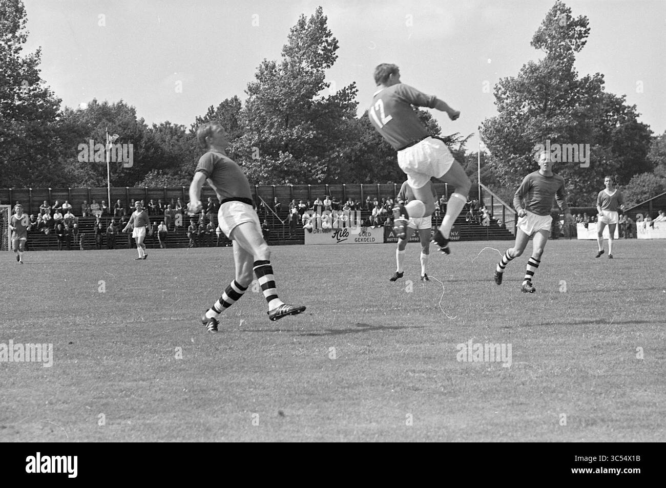 R.C.H. - Blue and White, Football, 08-08-1965 Whizgle News, Dutch Desk, Paesi Bassi, 1950-2000 è in corso una partita di calcio con giocatori in azione su un campo erboso. Un giocatore, con il numero 12, salta abilmente per colpire la palla, mentre due compagni di squadra e un avversario osservano il gioco. Gli spettatori possono essere visti seduti sullo sfondo, guardando con attenzione. Foto Stock