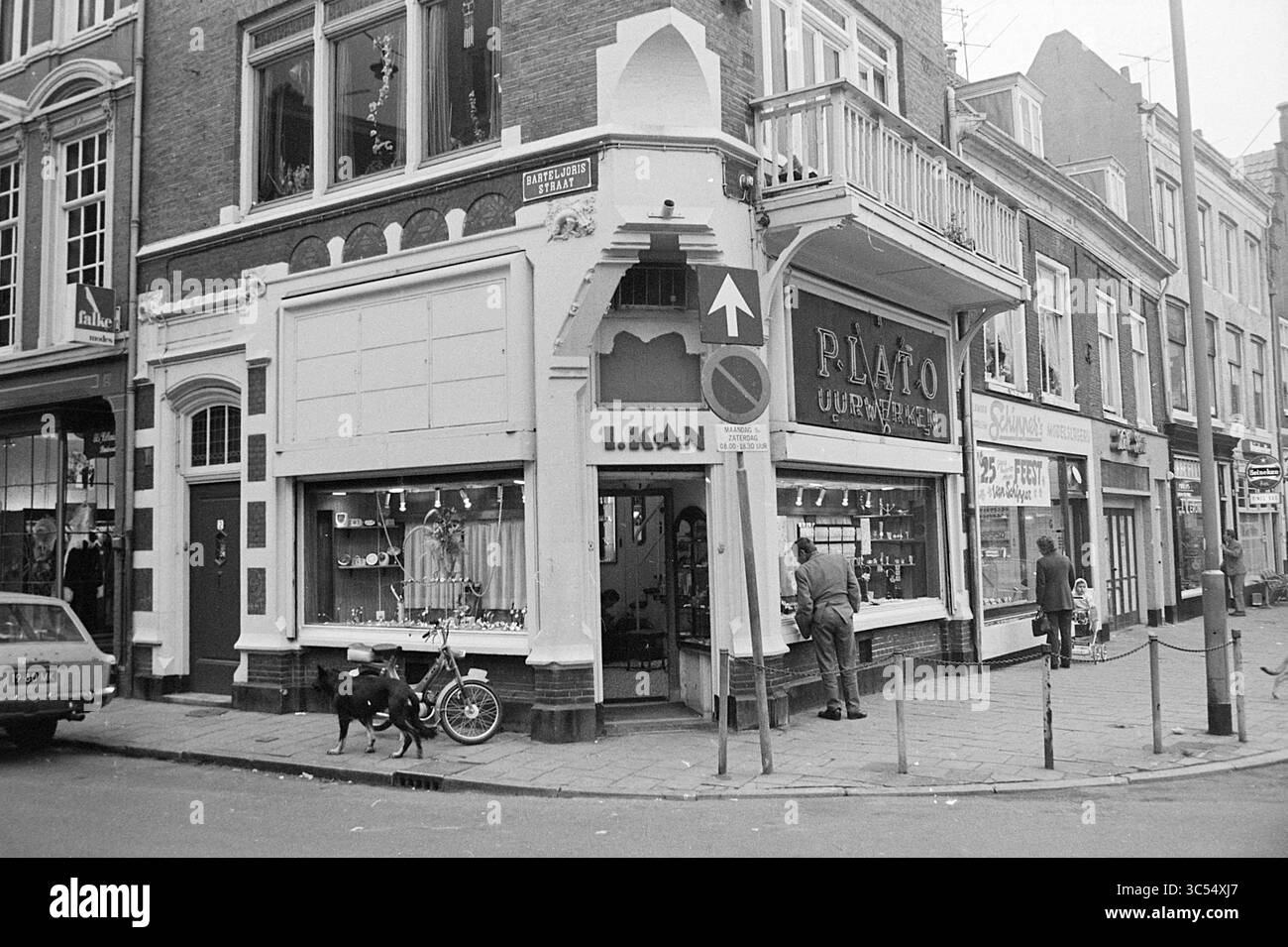 -0, Haarlem, Nederland Whizgle News, Dutch Desk, Paesi Bassi, 1950-2000 Una pittoresca scena di strada presenta un negozio all'angolo con dettagli architettonici ornati, che presenta una varietà di oggetti nelle sue vetrine. Una bicicletta si appoggia contro l'edificio, mentre un cane rimane nelle vicinanze. Le persone sono viste interagire con le vetrine, creando un'atmosfera vivace nel quartiere. Foto Stock