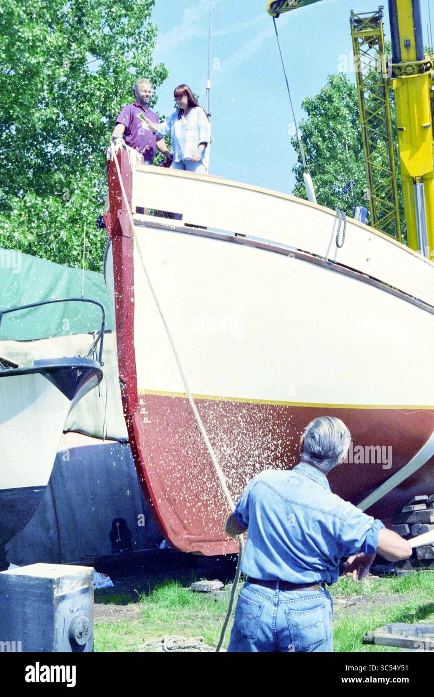 Varo delle barche, 26-06-1995 Whizgle News, Dutch Desk, Paesi Bassi, 1950-2000 due persone si trovano sul ponte di una barca, osservando come un'altra persona lavora nelle vicinanze, gestendo una corda. La scena cattura un momento di collaborazione in un cantiere, circondato dal verde e da altre imbarcazioni. Foto Stock