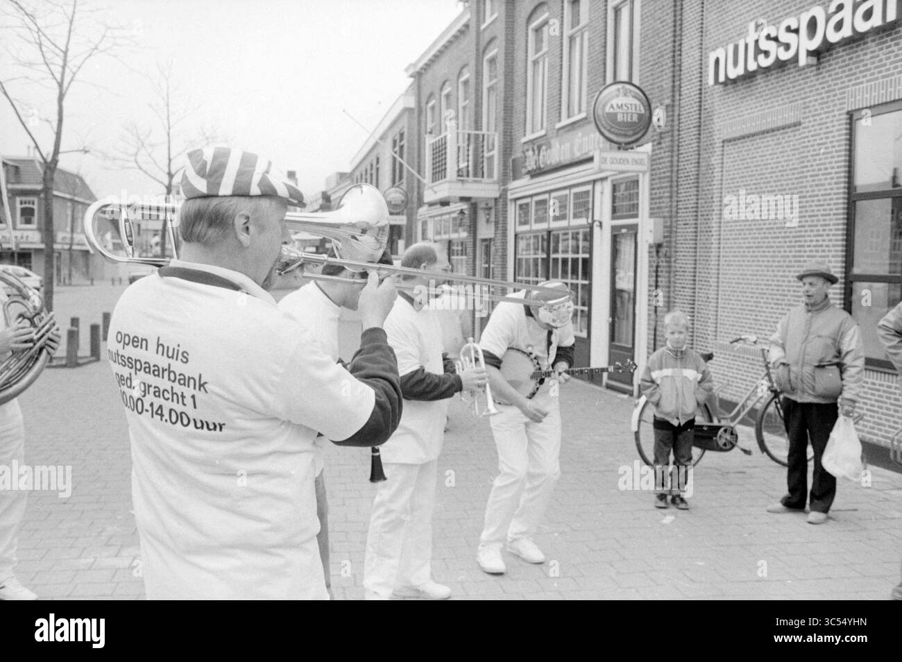 Open Day Office Schagen, Nuts, United Savings Banks, Schagen, 09-04-1988 Whizgle News, Dutch Desk, Paesi Bassi, 1950-2000 Un vivace spettacolo di strada con musicisti che suonano strumenti di ottone, mentre una piccola folla, tra cui bambini e adulti, guarda e gode dell'atmosfera festosa. Alcuni musicisti indossano abiti bianchi con cappelli distintivi, che si aggiungono alla scena celebrativa vicino a una banca locale. Foto Stock