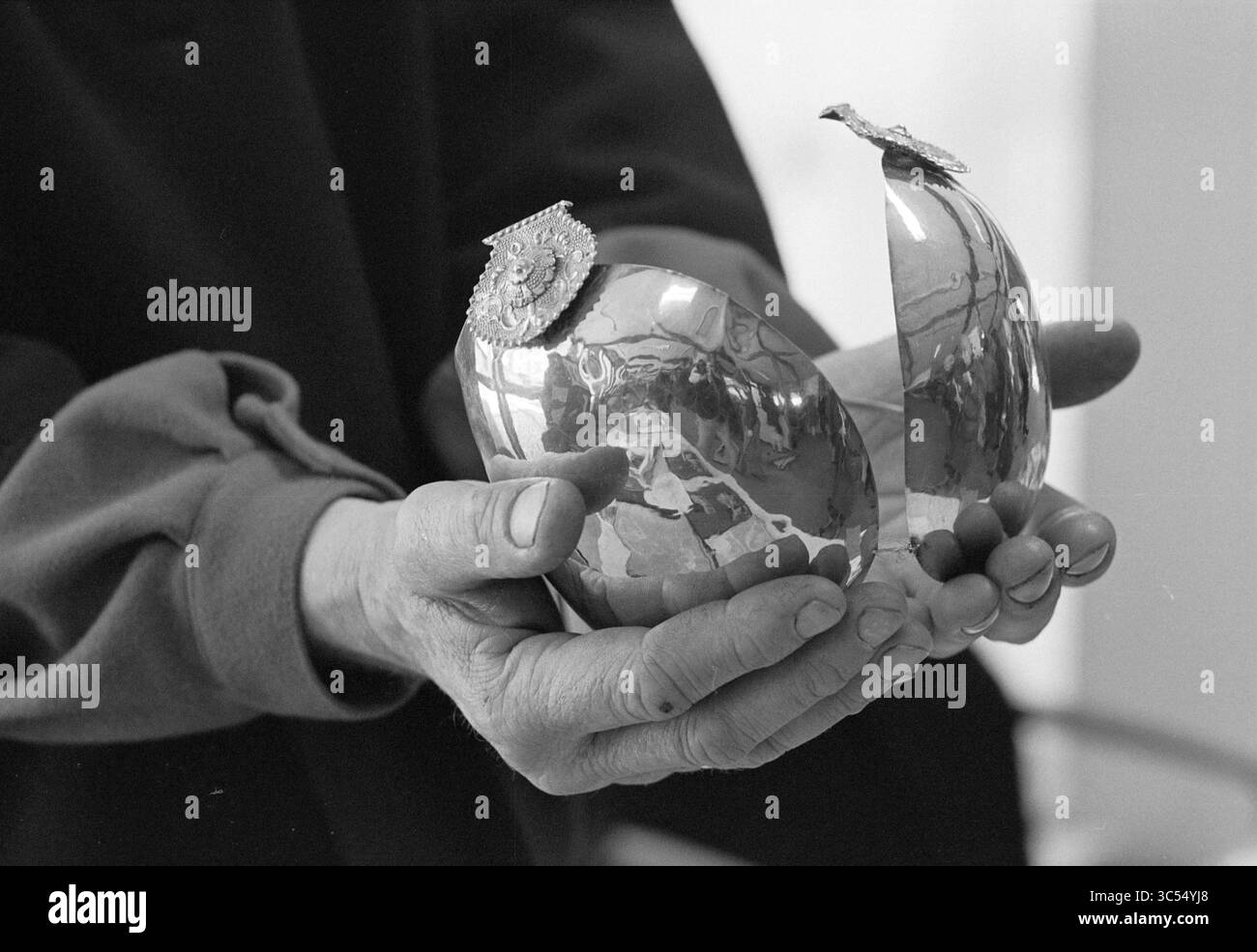Un cappuccio dorato sullo storico Market Whizgle News, Dutch Desk, Paesi Bassi, 1950-2000 Un paio di mani tiene con cura un oggetto a forma di uovo rotto splendidamente realizzato, decorato con intricati dettagli sulla parte superiore. La superficie riflettente cattura l'ambiente circostante, suggerendo il significato e l'arte dell'oggetto. Foto Stock