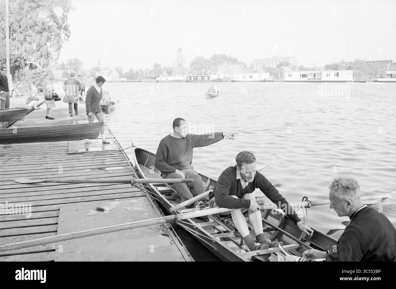 Medici durante le competizioni di canottaggio, canottaggio, canottaggio, ecc., 09-10-1965 Whizgle News, Dutch Desk, Paesi Bassi, 1950-2000 Un gruppo di uomini si prepara a remare, con uno che punta verso l'acqua mentre altri regolano le loro attrezzature. Nelle vicinanze, altri individui si impegnano in varie attività lungo il lungomare, creando una vivace scena sul fiume. Foto Stock