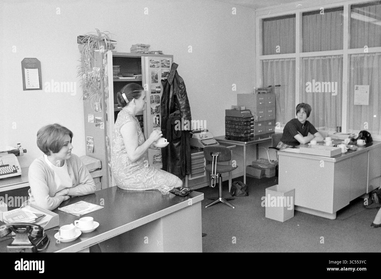 Tre signore in un ufficio Whizgle News, Dutch Desk, Paesi Bassi, 1950-2000 Un ambiente accogliente che ospita tre donne impegnate in varie attività. Una donna si siede a una scrivania e organizza i documenti, mentre un'altra, vestita elegantemente, si gode una tazza di caffè seduta su un armadietto. La terza donna si rilassa nelle vicinanze, contribuendo a creare un'atmosfera collaborativa in uno spazio di lavoro pieno di documenti e tocchi personali. Foto Stock