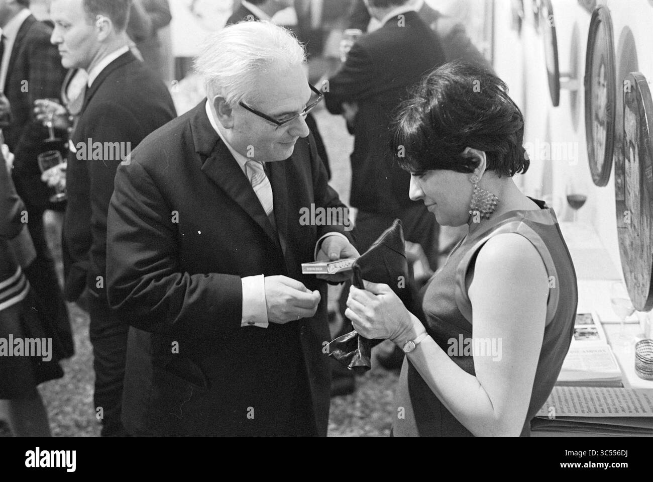 Apertura della Gallery T, Exhibitions, 07-04-1967 Whizgle News, Dutch Desk, Paesi Bassi, 1950-2000 un uomo e una donna si impegnano in una vivace conversazione ad un incontro sociale, con la donna che tiene in mano un piccolo oggetto mentre l'uomo sembra presentarle qualcosa. Lo sfondo è pieno di persone che si mescolano, suggerendo un evento vivace ed elegante. Foto Stock