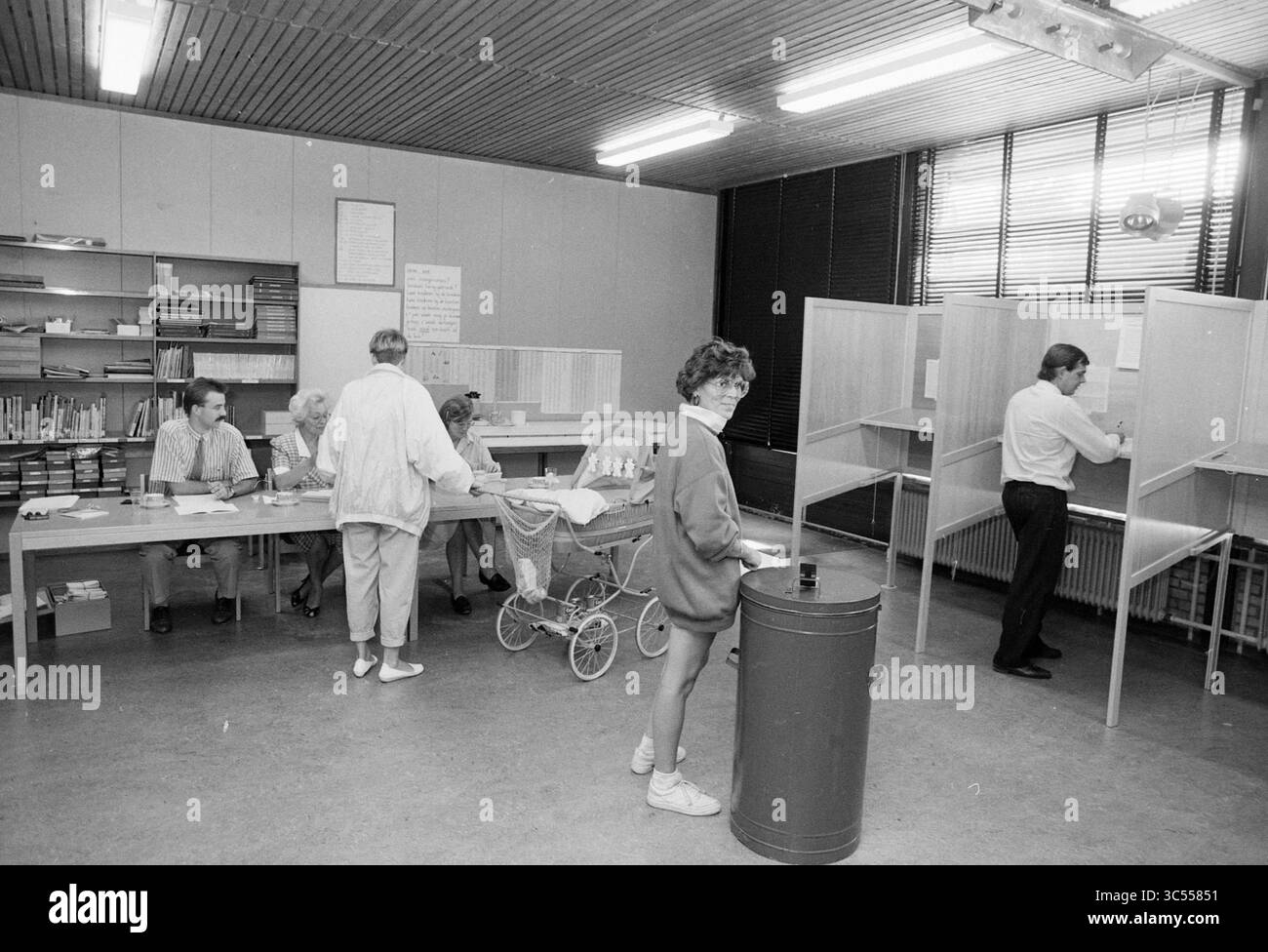 Sede elettorale Hillegom, in School, Hillegom, 06-09-1989 Whizgle News, Dutch Desk, i Paesi Bassi, 1950-2000 Un gruppo di persone è impegnato in varie attività in una sala progettata per la partecipazione civica, con diverse persone ai tavoli che elaborano documenti e altri che utilizzano cabine private, mentre un bambino in un passeggino è presente nelle vicinanze. L'ambiente presenta scaffali con libri e una bacheca sul muro. Foto Stock