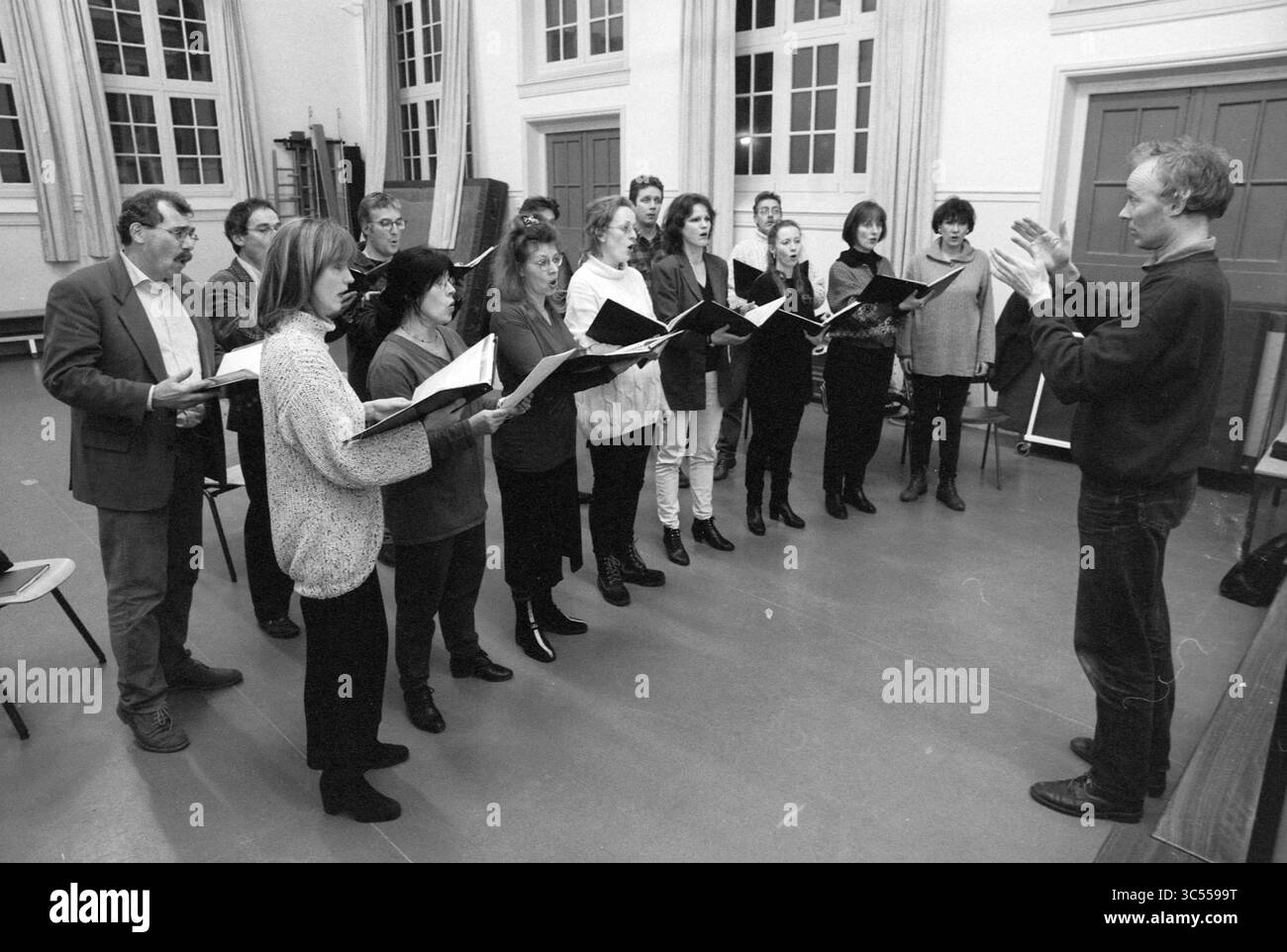 Prove del coro, Les Moucherons, 26-01-1997 Whizgle News, Dutch Desk, Paesi Bassi, 1950-2000 Un gruppo di membri del coro si trova in formazione, tenendo spartiti mentre seguono attentamente il direttore, che gesta espressivamente di fronte a loro. L'ambiente presenta grandi finestre ed elementi in legno che esaltano l'atmosfera di una sessione pratica. Foto Stock