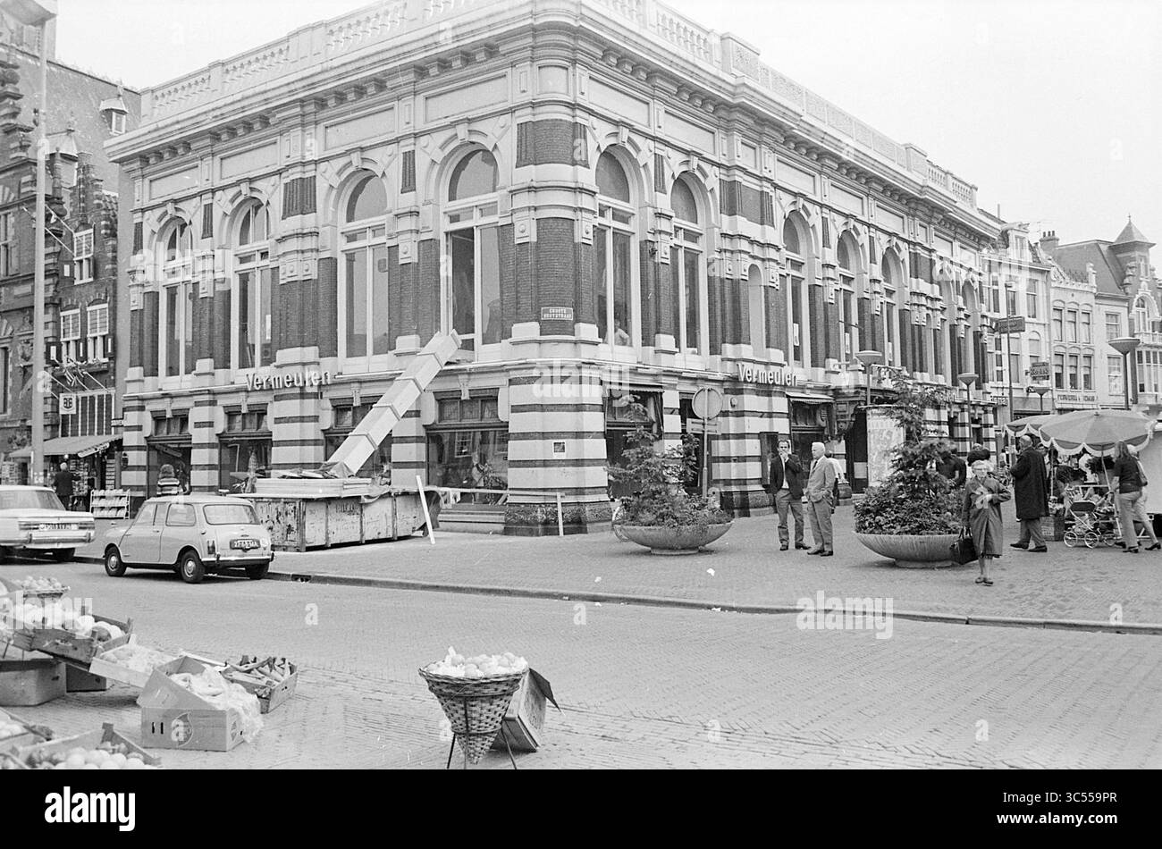 Esterno Vermeulen Grote Markt, Exterior, Haarlem, Grote Markt, Paesi Bassi, 10-08-1971 Whizgle News, Dutch Desk, The Netherlands, 1950-2000 Un vivace angolo di strada presenta un edificio ornato con grandi archi, circondato da venditori ambulanti che vendono merci. Le persone sono impegnate in varie attività, mentre le auto d'epoca sono parcheggiate nelle vicinanze. Le fioriere decorative aggiungono verde alla scena, catturando un'atmosfera urbana vivace. Foto Stock
