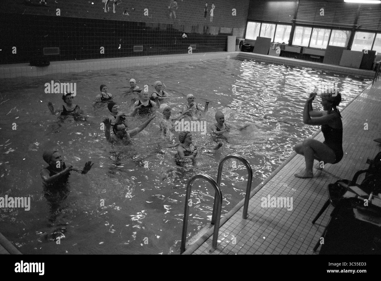 Swimming Whizgle News, Dutch Desk, The Netherlands, 1950-2000 Un gruppo di nuotatori si impegna in una routine sincronizzata in una piscina, mentre un istruttore si trova al bordo, guidandoli con segnali manuali. L'atmosfera e' vivace mentre i nuotatori sprigionano e tifanno, creando un senso di lavoro di squadra e gioia nell'acqua. Foto Stock