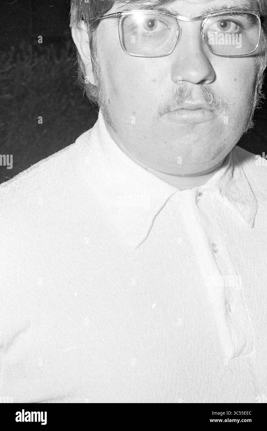 Ritratto dell'uomo con occhiali e baffi Whizgle News, Dutch Desk, Paesi Bassi, 1950-2000 Un giovane con occhiali si staglia con un'espressione seria, indossando una camicia con colletto. Ha capelli di colore chiaro e baffi, e lo sfondo presenta fogliame sfocato. Foto Stock