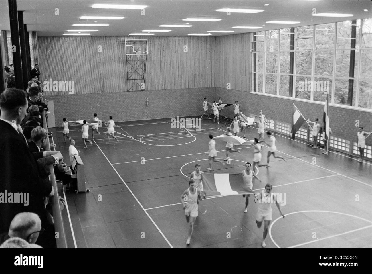 Sfilata di bandiere in una palestra Whizgle News, Dutch Desk, Paesi Bassi, 1950-2000 un gruppo di atleti si sta impegnando in un vivace spettacolo, sventolando bandiere e correndo su un pavimento di palestra. Gli spettatori guardano da un'area sopraelevata, mentre le grandi finestre consentono alla luce naturale di illuminare lo spazio. Le pareti in legno e l'alto soffitto della palestra creano un'atmosfera spaziosa per l'evento. Foto Stock
