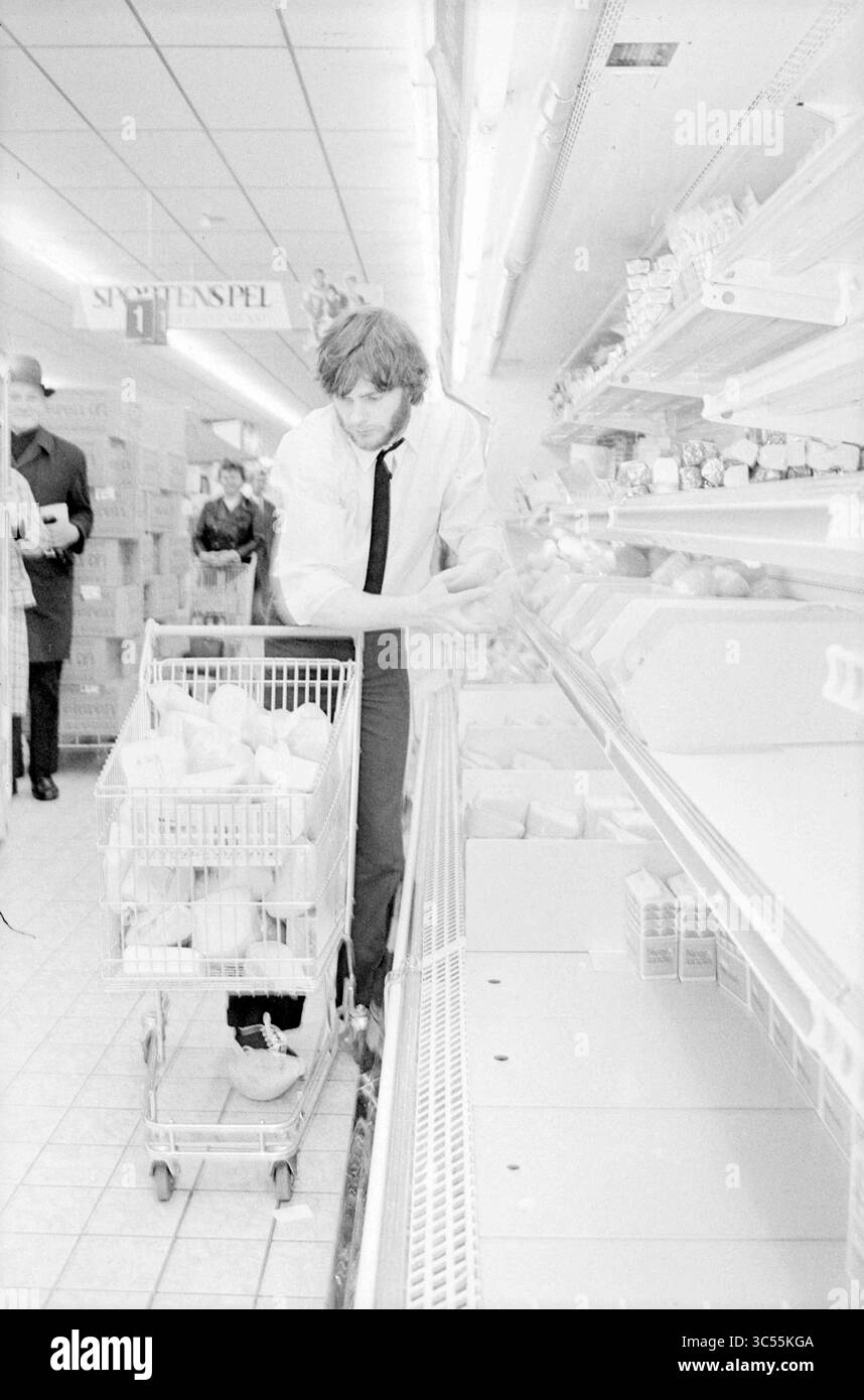 Shopping gratuito Albert Heijn, prezzi, 04-05-1972 Whizgle News, Dutch Desk, Paesi Bassi, 1950-2000 Un uomo con una camicia e una cravatta organizza con cura gli oggetti su uno scaffale mentre spinge un carrello attraverso un corridoio di un negozio di alimentari, con gli acquirenti sullo sfondo che navigano sugli scaffali. Foto Stock