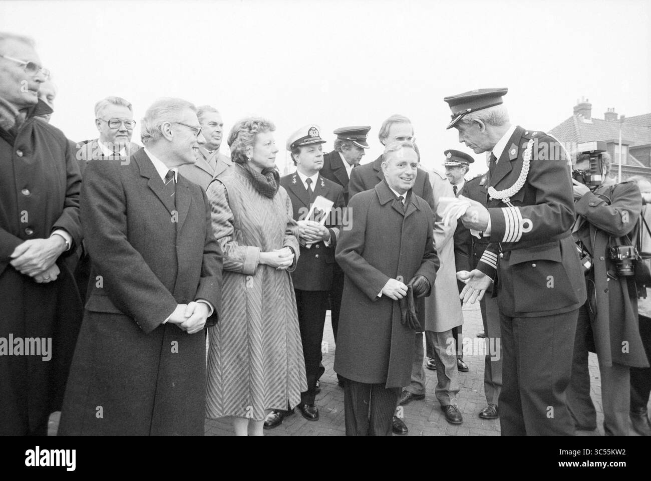 Installazione della guardia costiera nazionale a IJmuiden, installazione, guardia costiera, IJmuiden, Paesi Bassi, 26-02-1987 Whizgle News, Dutch Desk, Paesi Bassi, 1950-2000 Un gruppo di dignitari e funzionari si impegnano in conversazione, con una figura in uniforme che sembra presentare qualcosa ad un'altra. L'atmosfera riflette un evento formale, evidenziando le interazioni tra i partecipanti vestiti con abiti e cappotti vari. Foto Stock