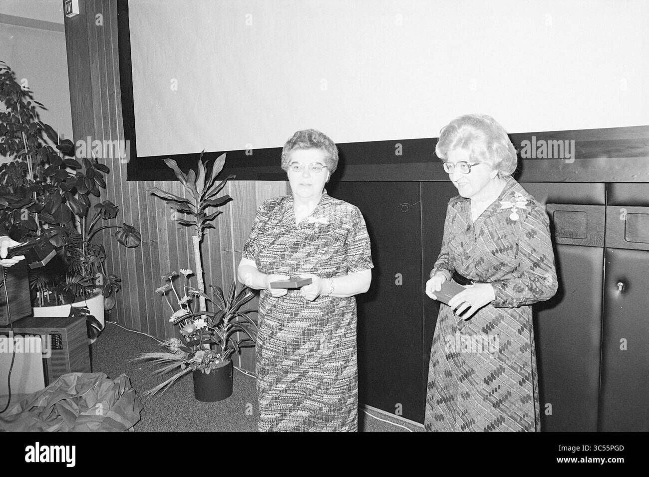 Premi Zeeweg Hospital IJmuiden, Awards, 12-05-1977 Whizgle News, Dutch Desk, Paesi Bassi, 1950-2000 due donne stanno di fronte a un grande schermo, interagendo l'una con l'altra. Sono vestiti con abiti a motivi geometrici e sembrano essere in un ambiente formale, forse durante una presentazione o un evento. Le piante in vaso sono visibili sullo sfondo, contribuendo all'atmosfera della scena. Foto Stock
