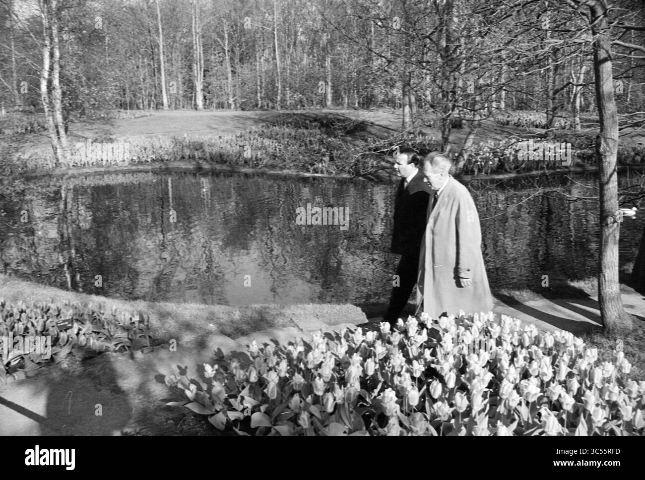 Il principe orientale visita Keukenhof, la tenuta della Fondazione Keukenhof a Lisse, 02-05-1963 Whizgle News, Dutch Desk, Paesi Bassi, 1950-2000 due persone passeggiano lungo un tranquillo stagno, circondato da un paesaggio di alberi e tulipani in fiore, suggerendo un momento di riflessione in un ambiente tranquillo. Foto Stock