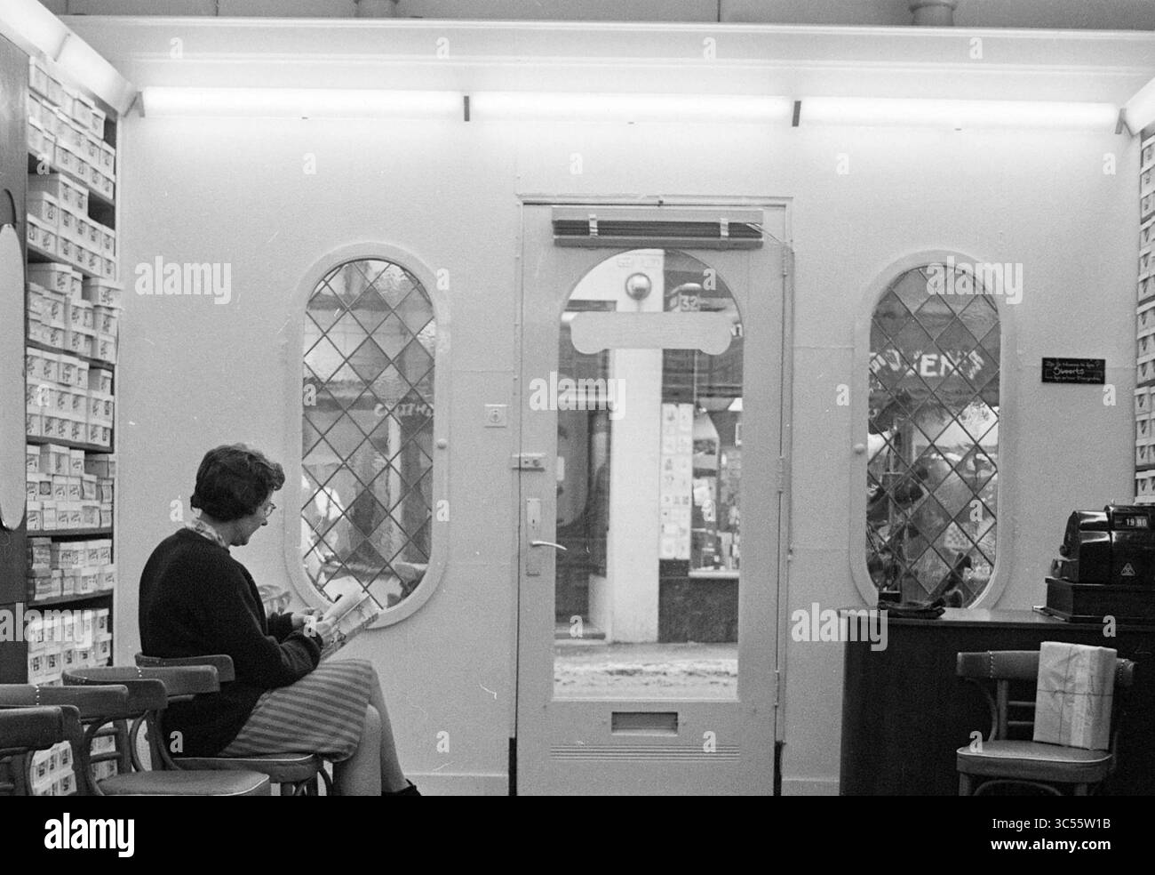 Silenzio nei negozi, negozi e chioschi, 12-02-1963 Whizgle News, Dutch Desk, Paesi Bassi, 1950-2000 Una donna siede tranquillamente in un negozio accogliente, la sua attenzione si concentra su un libro. L'entrata presenta eleganti finestre a specchio che offrono uno scorcio della strada all'esterno. L'atmosfera è tranquilla e invitante, incorniciata da scaffali pieni di articoli ordinati. Foto Stock